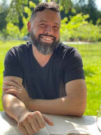 a bearded man smiling at a table with a glass of wine